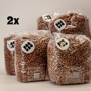 May include: Five clear plastic bags filled with brown grains, arranged in a group. Each bag has a white circular sticker with a black logo. The number "2x" is printed in black in the upper left corner.