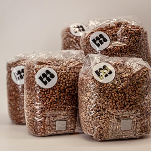 Könnte beinhalten: Mehrere transparente Plastiktüten, gefüllt mit braunen Körnern, sind vor einem weißen Hintergrund gestapelt. Jede Tüte hat einen weißen runden Aufkleber mit einem schwarz-grünen Logo. Die Tüten sind versiegelt und scheinen einsatzbereit zu sein.