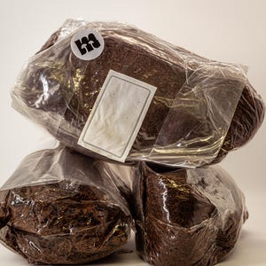 May include: Three brown, rectangular blocks wrapped in clear plastic. Each block is sealed with clear tape. A white label with a gray border is affixed to the top block. A white circular sticker with a black logo is also on the top block.