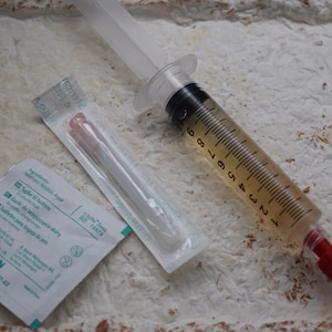 Op de afbeelding: Een doorzichtige plastic injectiespuit gevuld met een licht amberkleurige vloeistof, voorzien van een rode dop en zwarte maatstreepjes. Daarnaast liggen een steriele naald in een plastic verpakking en een B. Braun huidreinigingsdoekje.