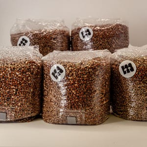 May include: Five clear plastic bags filled with brown grains. Each bag has a white circular sticker with a black logo. The bags are arranged on a white surface.