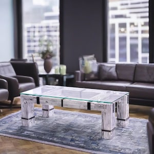 May include: A whitewashed wood coffee table with a glass top sits in a modern living room. The table has a rustic, reclaimed wood look and features black metal accents. The table is surrounded by dark gray leather furniture and a gray rug.