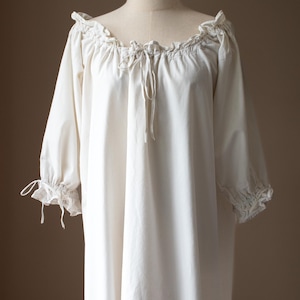 May include: An off-white, long-sleeved nightgown featuring a gathered, elasticized neckline with a central drawstring tie. The sleeves are three-quarter length with ruffled, elasticized cuffs that also include small, decorative drawstring ties.