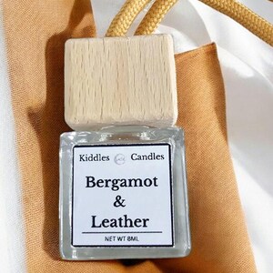 Puede incluir: Una botella de vidrio transparente con una tapa de madera y una cuerda dorada. La botella contiene un ambientador de coche con la etiqueta "Kiddle Candles Bergamot & Leather Net WT 8ML".
