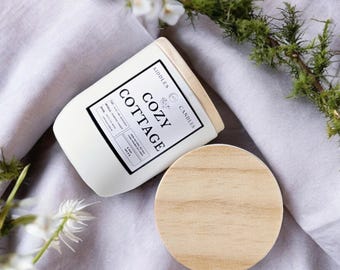 May include: White candle jar with a wooden lid. The label reads "Cozy Cottage" and "Kiddle's Candles".