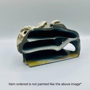 May include: A decorative, layered rock-like structure with three openings. The object is primarily gray with hints of yellow and white paint. The text at the bottom reads, "Item ordered is not painted like the above image*."