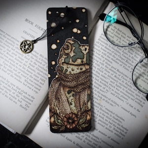 May include: A wooden bookmark with a hand-painted design of a frog wearing a scarf and surrounded by flowers. The background is black with white snowflakes.