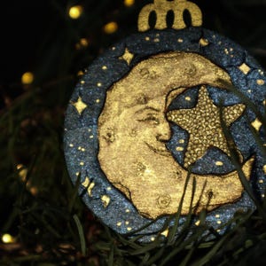 May include: A celestial-themed Christmas ornament featuring a gold crescent moon and star against a dark blue background with sparkling accents. The ornament is round with a decorative top and is hanging among green pine needles.