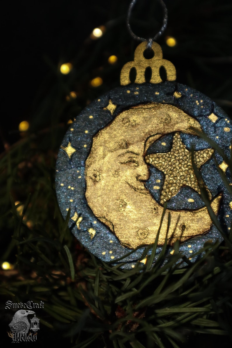 May include: A celestial-themed Christmas ornament featuring a gold crescent moon and star against a dark blue background with sparkling accents. The ornament is round with a decorative top and is hanging among green pine needles.