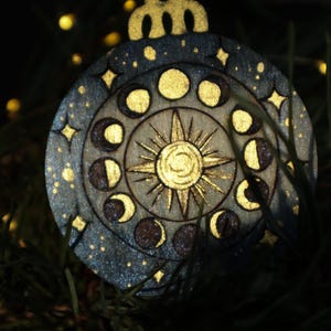 May include: Wooden celestial ornament with a gold sun, moon phases, and stars. The ornament is round with a decorative top and a silver string for hanging. The design is painted on a wooden surface with a dark blue and gold color scheme.