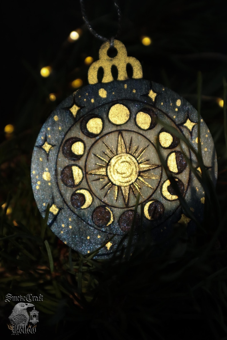 May include: Wooden celestial ornament with a gold sun, moon phases, and stars. The ornament is round with a decorative top and a silver string for hanging. The design is painted on a wooden surface with a dark blue and gold color scheme.