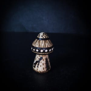 Könnte beinhalten: Eine kleine, helle Pilzfigur mit detaillierten Brandmalereien. Der Hut zeigt Alien-Gesichter und ein gepunktetes Band, während der Stiel die Silhouette einer Person in der Nähe eines Tannenbaums in einer Waldumgebung darstellt.