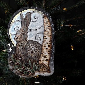 Peut inclure: Ornement de Noël en bois représentant un lièvre avec un flocon de neige sur la poitrine, sur fond de clair de lune. L'ornement comprend un bouleau et des brins de verdure, avec le texte "SmokeCraft Hollow".