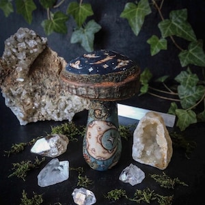 May include: A whimsical, hand-painted mushroom sculpture with a celestial design. The cap is dark blue with stars and a crescent moon, while the stem features a moon and star motif. Crystals and moss surround the mushroom, creating a mystical scene.