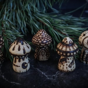 May include: Five hand-painted wooden mushroom figurines with intricate designs. Each mushroom features a unique pattern, including stars, moons, and geometric shapes. The mushrooms are painted in shades of brown and white.
