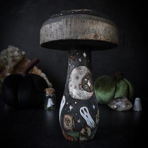 May include: A decorative wooden mushroom sculpture featuring a dark painted stem adorned with illustrations of a large moon, white ghosts, tombstones, and small stars. The mushroom cap has a natural wood grain texture and a dark painted top surface.