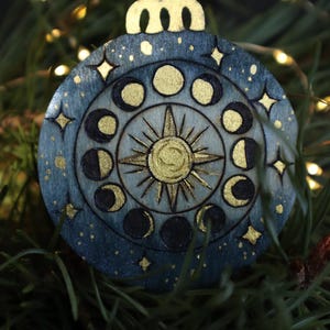 May include: A round, blue and gold celestial-themed Christmas ornament. The design features phases of the moon, stars, and a sunburst. The ornament is hanging from a silver string. The text "SmokeCraft Hollow" is visible at the bottom.
