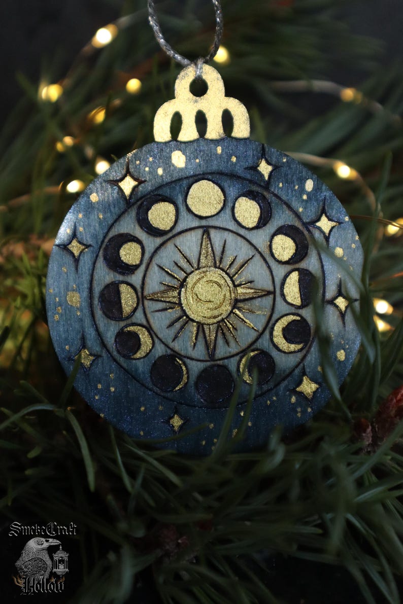May include: A round, blue and gold celestial-themed Christmas ornament. The design features phases of the moon, stars, and a sunburst. The ornament is hanging from a silver string. The text "SmokeCraft Hollow" is visible at the bottom.