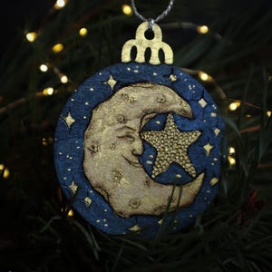 May include: A celestial-themed Christmas ornament. The round ornament features a smiling gold moon and a gold star on a dark blue background, with gold stars scattered around. It has a gold-colored top for hanging.