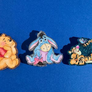 May include: Three glitter-covered character pins featuring Winnie the Pooh, Eeyore, and Tigger. The pins are all in a cartoon style and have a sparkly finish.