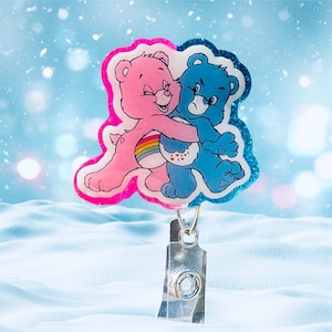 May include: A pink and blue Care Bear character badge with a rainbow and glitter accents. The badge is attached to a silver retractable badge holder.
