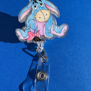 May include: A blue glitter badge holder featuring a cartoon image of Eeyore the donkey from Winnie the Pooh. Eeyore is sitting down with a sad expression and a pink bow on his tail.