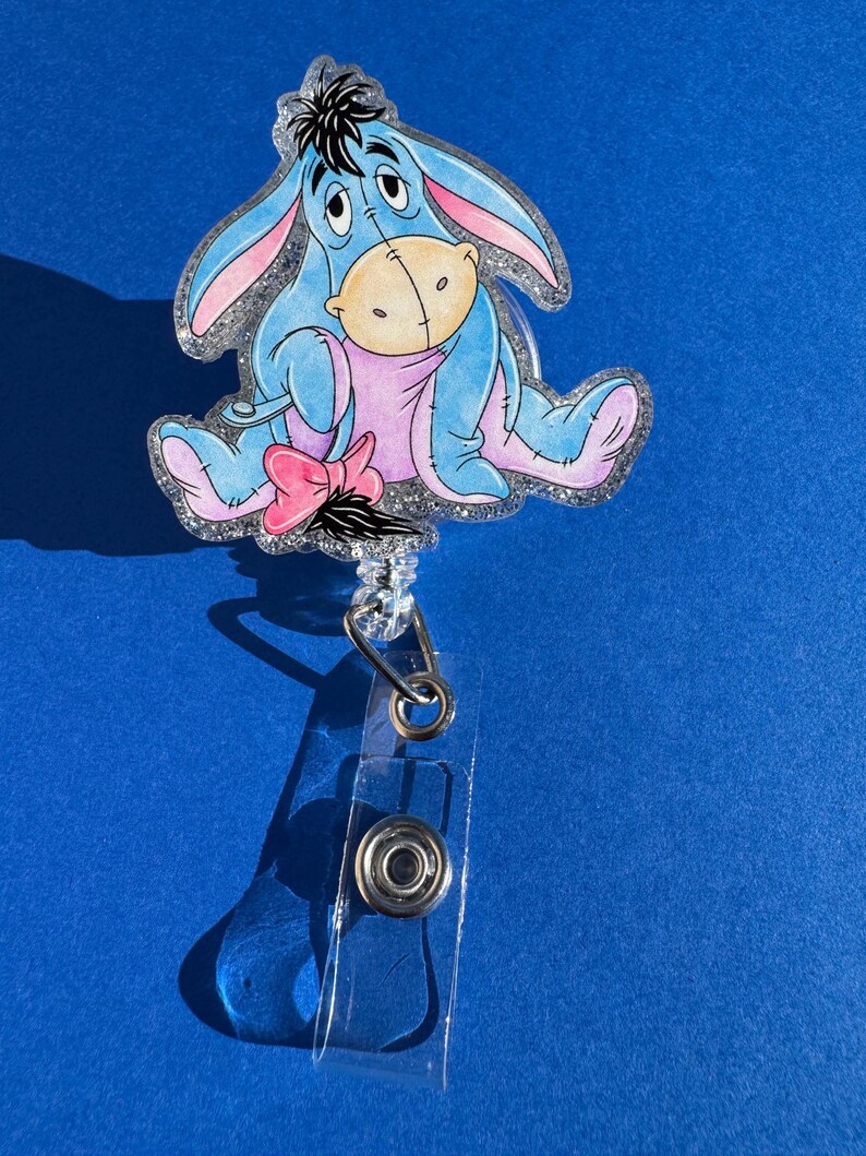 May include: A blue glitter badge holder featuring a cartoon image of Eeyore the donkey from Winnie the Pooh. Eeyore is sitting down with a sad expression and a pink bow on his tail.