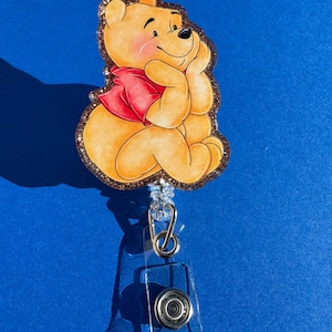 May include: A Winnie the Pooh character badge reel with a gold glitter background. The character is sitting with a red shirt and a thoughtful expression.