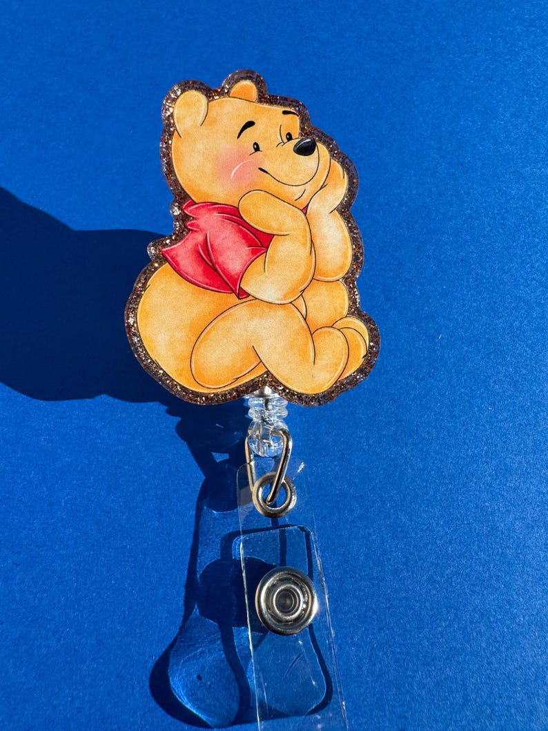 May include: A Winnie the Pooh character badge reel with a gold glitter background. The character is sitting with a red shirt and a thoughtful expression.