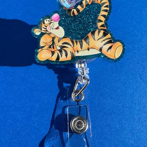 May include: A teal glitter badge holder featuring a cartoon image of Tigger from Winnie the Pooh. Tigger is lying on his back with his tail curled up and a butterfly above him.