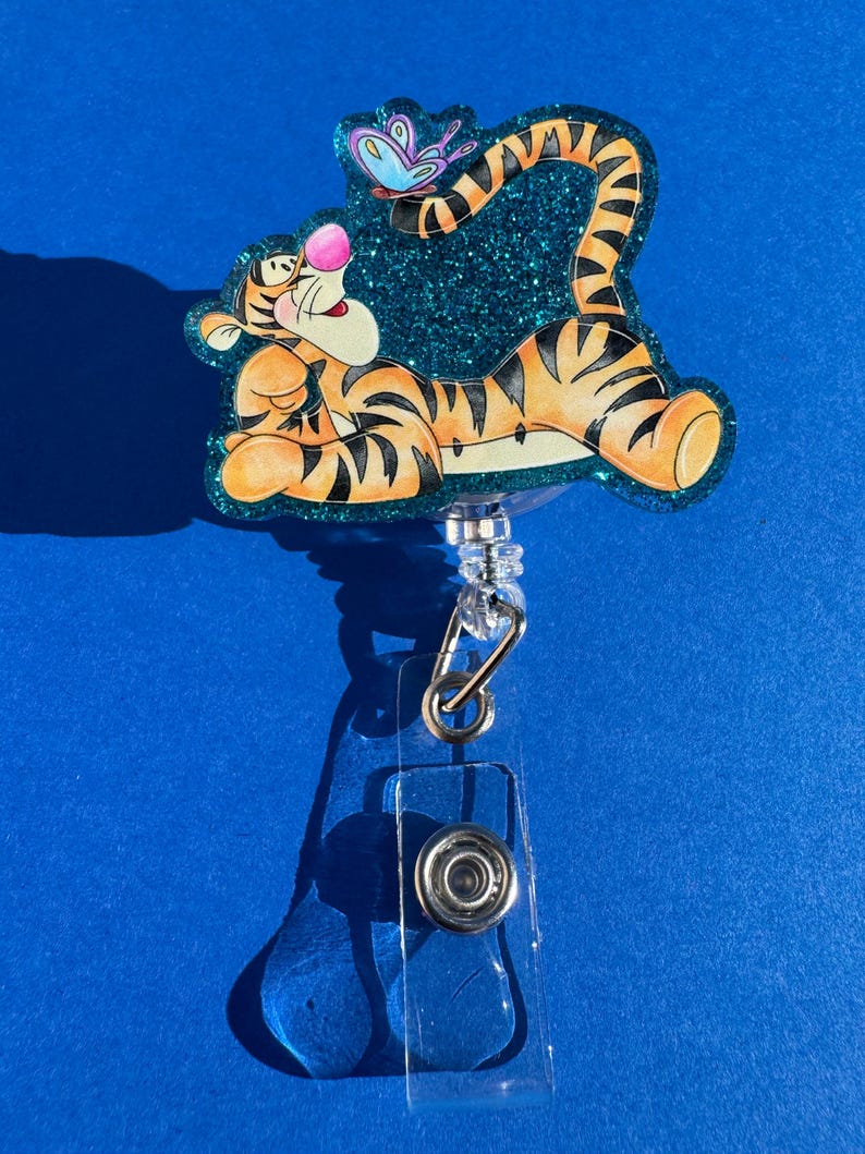 May include: A teal glitter badge holder featuring a cartoon image of Tigger from Winnie the Pooh. Tigger is lying on his back with his tail curled up and a butterfly above him.