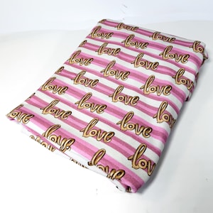 May include: Folded fabric with a repeating pattern of the word "love" in gold script over pink and white stripes. The fabric is folded neatly, showcasing the design.