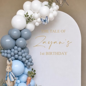 May include: A decorative arch for a first birthday celebration, featuring a balloon garland in white, blue, and beige. The arch includes Peter Rabbit and a duck cutout, with the text "The Tale of Zayan's 1st Birthday" in gold.