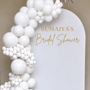 May include: A white arched backdrop with gold script reading "SUMAIYA'S Bridal Shower". A cascade of white balloons of various sizes decorates the left side, creating a celebratory and stylish display for a bridal shower.
