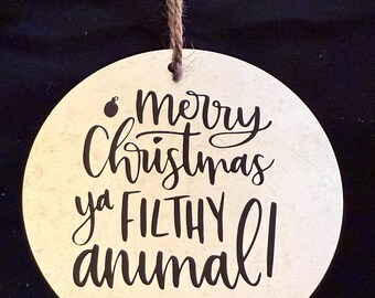 Op de afbeelding: Een ronde, beige houten ornament met een bruine touwhalter. De ornament heeft zwarte tekst die "merry Christmas ya FILTHY animal!" leest.