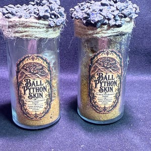 May include: Two clear glass jars filled with gold glitter, each topped with dark gray stones and wrapped in twine. The labels read: Ball Python Skin, For use in Transformation Potions, Promotes shedding of the former form & taking of another.