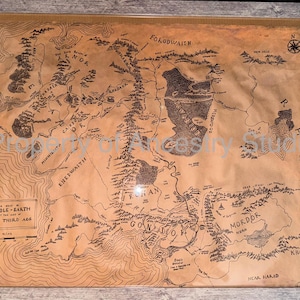 May include: A framed map of Middle-earth from The Lord of the Rings, rendered in sepia tones. The map details geographical features, with text labels and a compass rose. The frame is a light brown color.