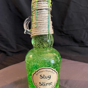 May include: A green glass bottle filled with a bubbly, green liquid labeled "Slug Slime." The bottle is sealed with a rope-wrapped top and green wax drips. A small handle is attached to the side. The label reads "Secretions to protect from those out to get you."