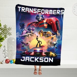 May include: A Transformers themed fleece blanket with a blue, red, and yellow color scheme. The blanket features a group of Transformers characters, including Optimus Prime, Bumblebee, and others. The text "TRANSFORMERS" is printed in white at the top of the blanket. The name "JACKSON" is printed in white at the bottom of the blanket.