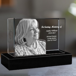 May include: A black and white photo of a woman is engraved in a clear glass cube on a black base. The text "In loving Memory of Sophia Williams 1958-2025 Devoted Teacher, Loving Mother, Sister & Friend" is engraved on the glass cube.