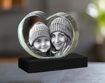 Puede incluir: Un pisapapeles de cristal en forma de corazón con una base de madera negra. El cristal presenta una foto en blanco y negro de una mujer y un niño con gorros de invierno.