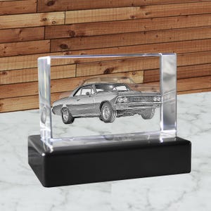 May include: A clear, rectangular crystal block with a detailed etched image of a classic car. The car is silver and sits on a black base. The background is a wooden wall.