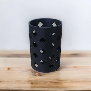 May include: A cylindrical, dark blue container with a diamond-shaped pattern cut out of the sides. The container is made of a solid material and sits on a light-colored wooden surface. The background is a plain white wall.