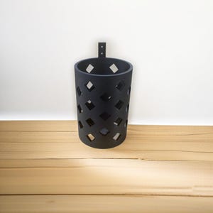 May include: A black metal wall-mounted storage container with a diamond pattern design. The container is empty and has a hook on the top.