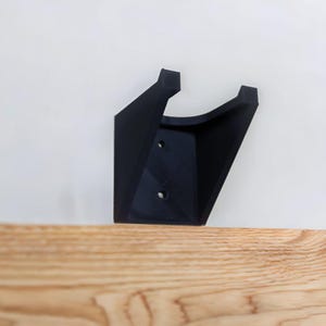 Op de afbeelding: Een zwarte plastic wandbeugel met twee schroefgaten. De beugel is ontworpen om een plank of ander object te dragen.