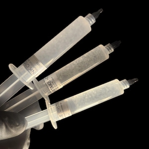 May include: Three clear syringes with black tips are held against a black background. Each syringe has a white label with black text. The syringes are filled with a cloudy liquid. The syringes are held by a gloved hand.