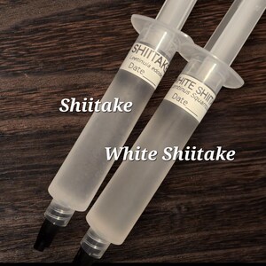 May include: Two clear syringes filled with a translucent liquid, labeled "Shiitake" and "White Shiitake." The syringes are angled against a dark wood grain background. Each syringe has a label with the text "Date".