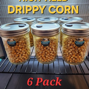 May include: Six glass jars filled with yellow kernels, labeled "High Yield Drippy Corn." Each jar has a metal lid and a small sticker. The jars are arranged on a black wire rack, with the text "6 Pack" in red.