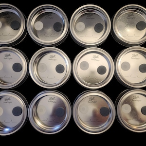May include: A collection of twelve Ball SureTight canning jar lids. Each lid is silver-colored and features a white and a black circular insert. The lids are arranged in three rows of four, against a black background.