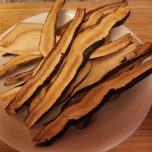 May include: A white plate filled with thin, dried, elongated slices of mushrooms. The slices feature a light tan interior with dark brown, textured edges, showing a fibrous and dehydrated appearance throughout the collection of pieces.